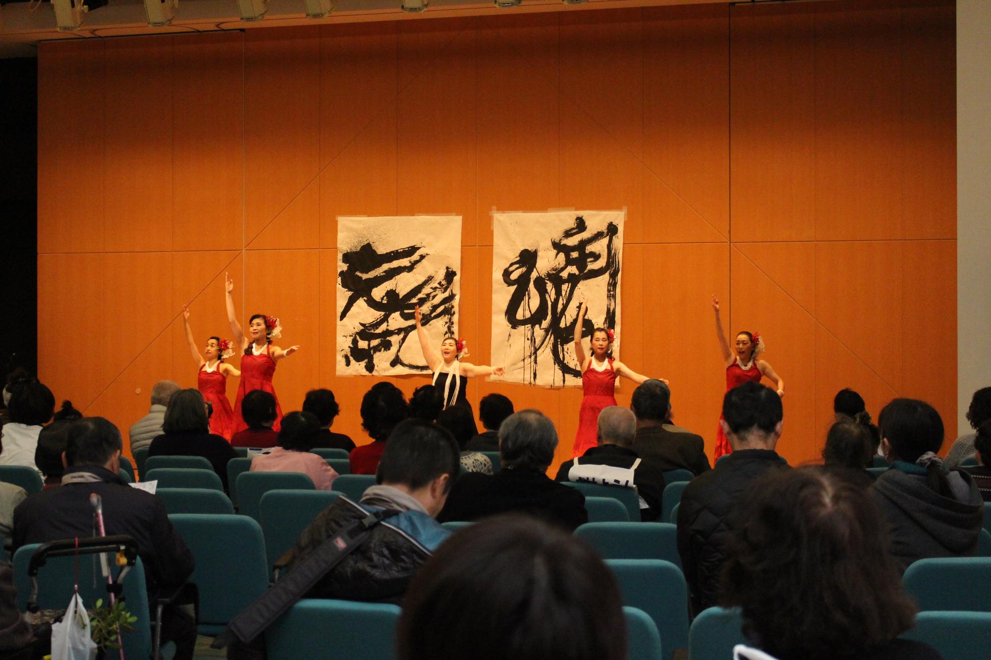 右手を上にあげ、左手を横に伸ばし、フラダンスを踊る5人の女性と、それを見ている人々の写真