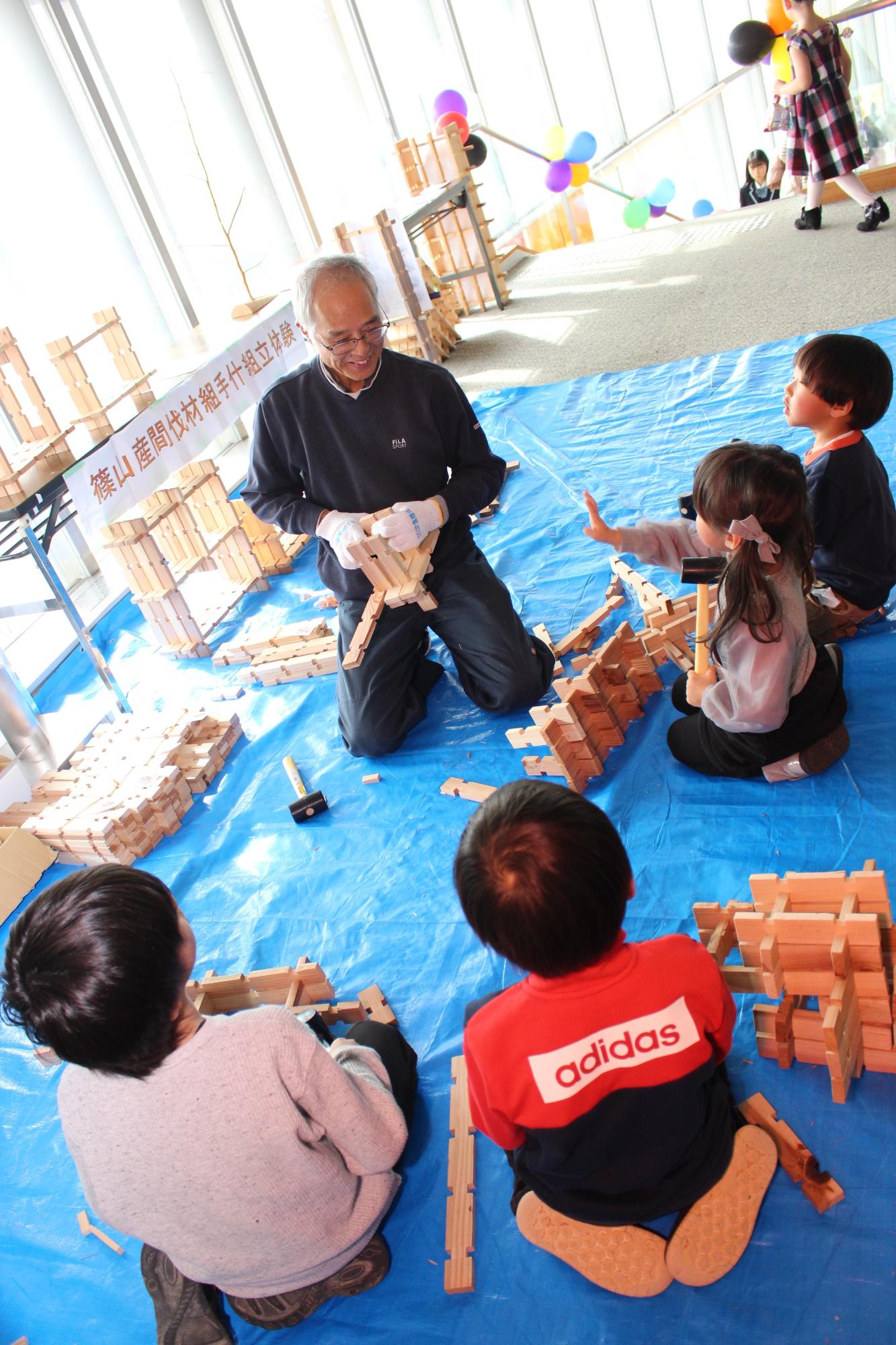 ブルーシートがひかれたところで篠山産の木材を使って木材を組む体験をする子どもたちと、それを教える男性の写真