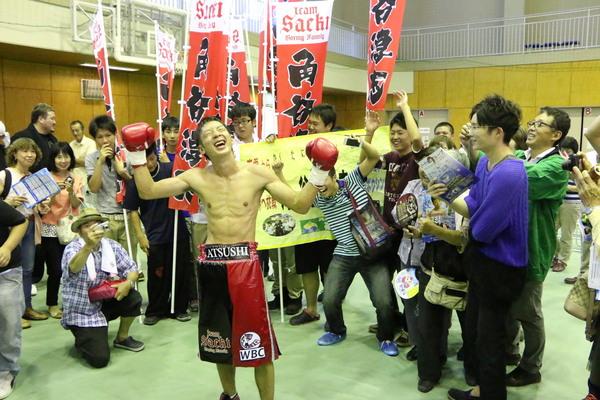 両手をあげ笑顔でガッツポーズをしている角谷選手を囲んで盛り上がっている応援団の方の写真