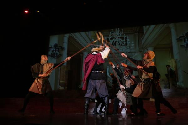 茶色のベレー帽を被り、赤いマントをしている男性に槍を向けている方々の写真