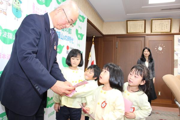 市長がさくら色の招待状を読んでおり、黄色いスモッグを着た園児4名のうちの1人が市長の顔を見ながら話をしている様子の写真