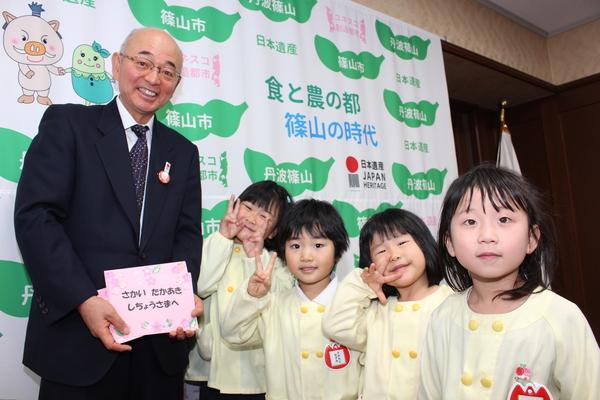 市長が「さかい たかあきしちょうさまへ」と書かれているさくら色の招待状を持って、園児達はピースサインをして笑顔で写っている写真