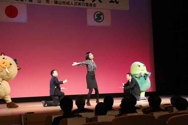 舞台の真ん中でストライプシャツにタイトスカート姿の女性が真ん中で手を広げて両脇にスーツ姿の男性が座って女性を盛り立てている様子をゆるキャラ2名が盛り上げている写真