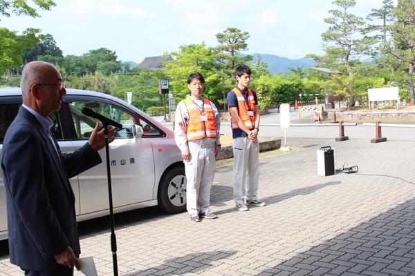 白い自動車の前に山本 圭太係長と伊藤 琢郎主事が立っており、市長がスタンドマイクの前で話をしている様子の写真