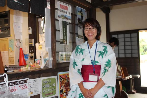 デカンショ館で眼鏡をかけた女性職員の方が白地に緑の模様に赤の帯の浴衣を着て笑顔で正面姿の写真