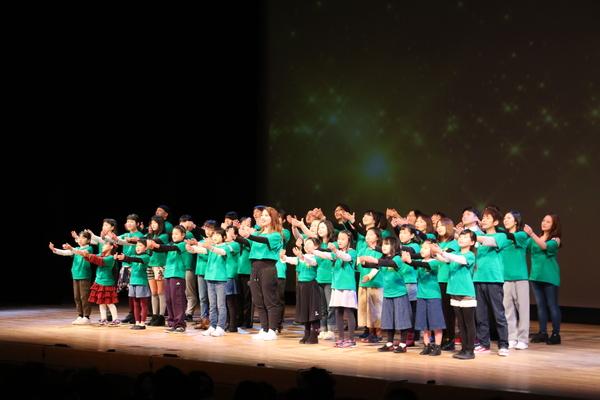 舞台で煌めくスクリーンの映像をバックに緑のTシャツを着たミュージカルの劇団が手を上げて何か表現している写真