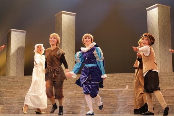 紫と水色の衣装を着た女性と茶色の衣装を着た主人公役の女性が笑顔で手を繋ぎスキップしており、その周りでは歌を歌い踊っている出演者の写真