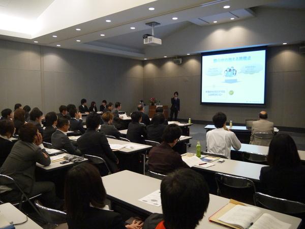 前方にあるスクリーンの映像を見ながらマイクを持った男性が説明をしており、参加者が椅子に座りテーブルには資料を広げ話を聞いている様子の写真