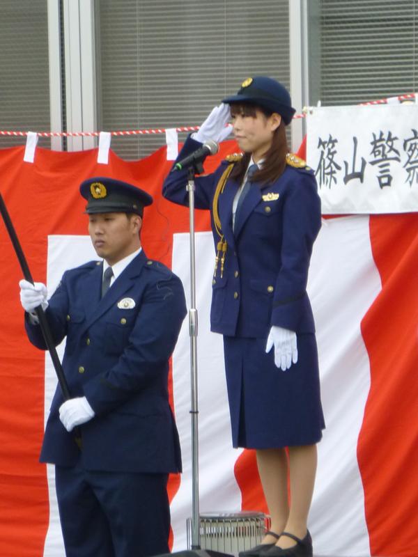 警官服姿の一日署長の竹内 沙也加さんが壇上で敬礼をし、その横で旗の棒を持って立っている警察官の写真