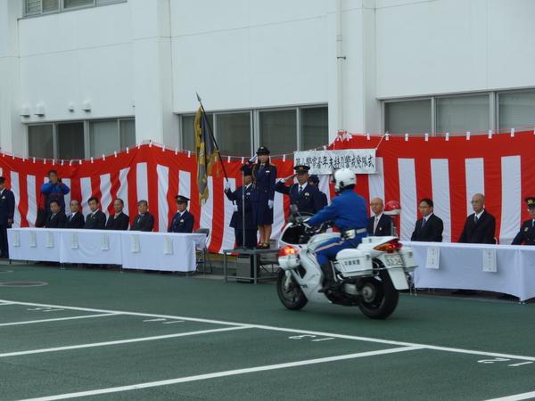 年末特別警戒発隊式で壇上に立つ警官服姿の竹内 沙也加さんや後ろにいる警察官が白バイ隊員に敬礼しているのを来賓者が見ている写真