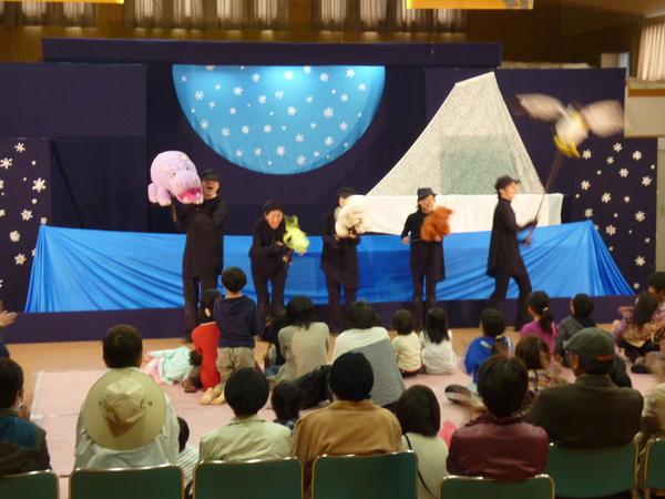 黒い衣装を着た5人の男女が人形を手に持ち劇をしている様子を真剣に子供達が見ている写真