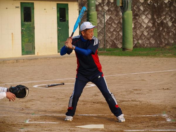 紺色ティーシャツを着て、白い帽子を被った前田部長がバッターボックスに立ち、ボールを打とうとバットを両手で握り、構えている様子の写真