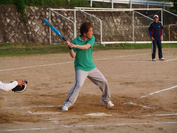 緑色のポロシャツを着た小山部長がバッターボックスに立ち、ボールを打とうとバットを両手で握り、構えている様子の写真