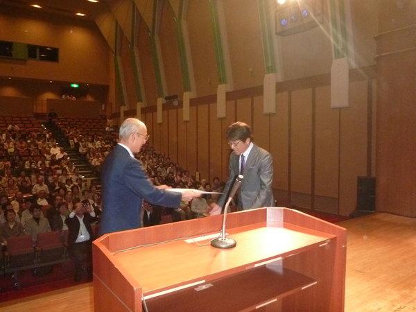 篠山市高齢者大学合同開講式の会場で、市長が丹波篠山ふるさと大使原田 伸郎さんに委嘱状を渡している写真