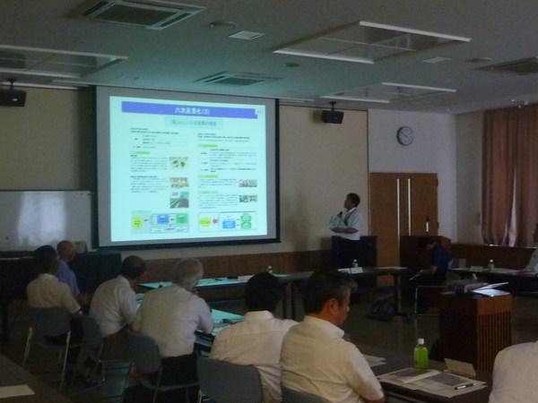 プロジェクターに映し出された映像を見ながら、マイクを持った男性が説明をしており、席についている出席者が映像を見ながら、真剣に話を聞いている様子の写真