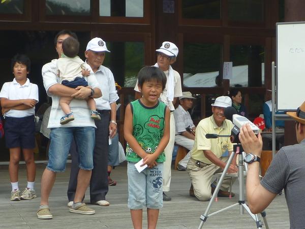 大声大会で緑のタンクトップに水色のズボン姿の男の子が大きい声をマイクにむかって出している写真
