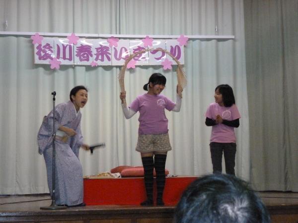 「後川春来いまつり」の出し物で女の子が割り箸を繋いだものを楕円形に丸めており、もう一人の女の子が拍手をしていて、司会者が声を上げている写真