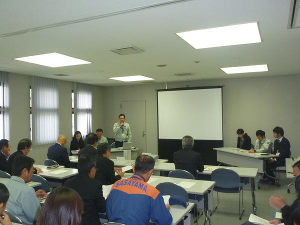 報告会で、スクリーンがあり、男性がマイクを持ち立って話をしている写真