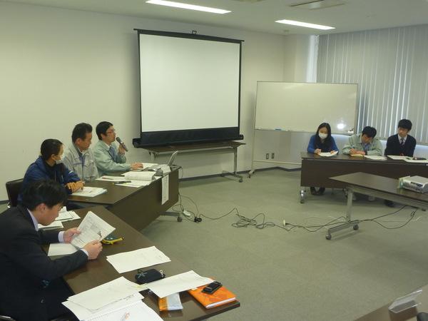 会議中にマイクを持った男性職員が話しているのを他の職員が資料を見ながら聞いている写真