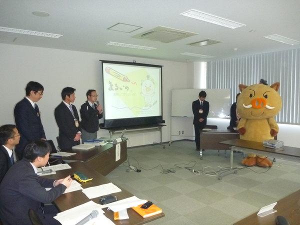 会議中、スクリーンにゆるキャラのイラストが映し出されており、マイクを持って発言している男性職員らとゆるキャラが立って聞いている写真