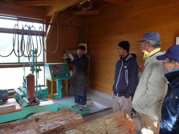 室内の中に手前には木材が置かれいる奥にはエメラルドグリーン色の大きな機械が置かれており、前掛けを付けている男性が説明しているのを参加者が聞いている様子の写真