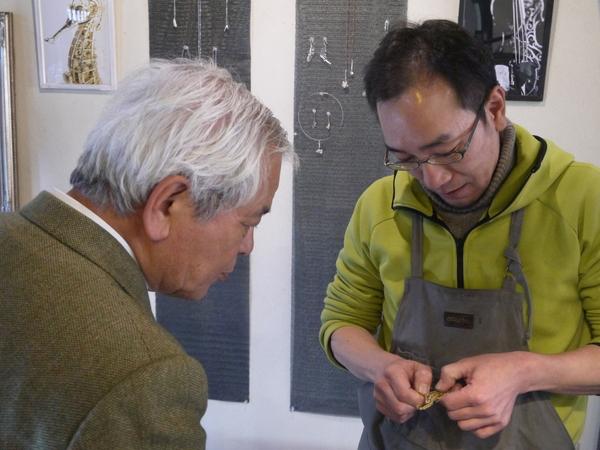 メガネをかけた男性が持っている應需細工をのぞいて説明を聞いている文化庁長官の写真