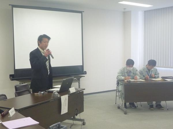 左前方の男性がマイクを持ち話をしていて、右後方の男性2名が長机に座り資料を見ている様子の写真