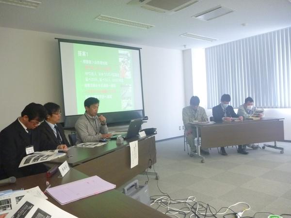 左前方の長机に男性3名が座っており、一番右に座っている男性がマイクを持ちスクリーンを使いながら説明していて、右前後方の長机には資料を見ている男性3名が座っている写真