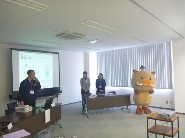 左前方で男性がマイクを持ち、スクリーンを使いながら説明していて、右横には両手を前で組み立っている女性2名とキャラクター（まるいの）の写真