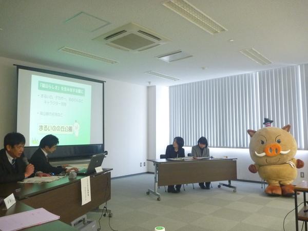 左前方の長机に男性2名が座っており、右に座っている男性がパソコンを使い、スクリーンに映し出し、左に座っている男性が資料を見ながら説明している。右後方の長机には女性2名が座っており、その横にキャラクター（まるいの）が座っている写真