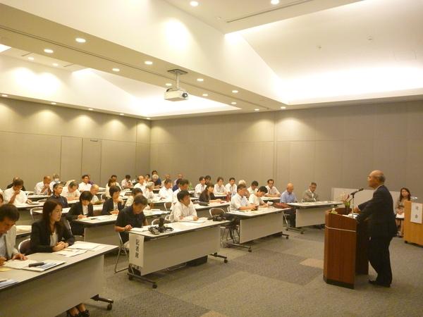 男性が演台の前にてマイクで話している内容を会場に来ている人たちが真剣に聞いている写真