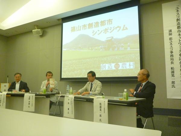 スクリーンの下で男性4名が座って討論している写真