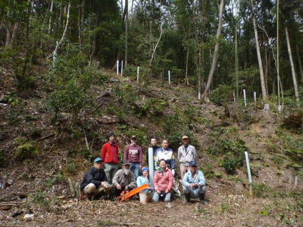 奥に木が茂っている森の中で9名の男性達と1名の女の子が並んでいる集合写真
