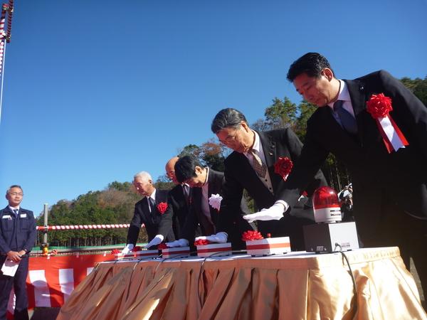 紅白のリボンが巻かれたボタンが机の上に5台設置されており、市長や関係者が一斉に白い手袋を付けた右手で、ボタンを押している様子の写真