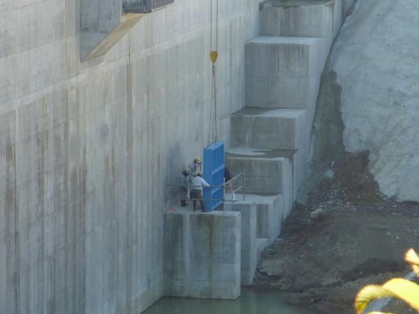 ダムの壁に階段のようなものが作られており、作業員が青い四角い取水ゲートをクレーンのロープで持ち上げているのを確認しながら補佐している様子の写真