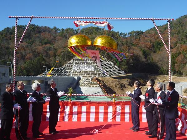 紅白の横断幕で覆われた舞台の上で、西紀ダム湛水式の金色のくす玉が割れて、関係者が手を叩いている様子の写真