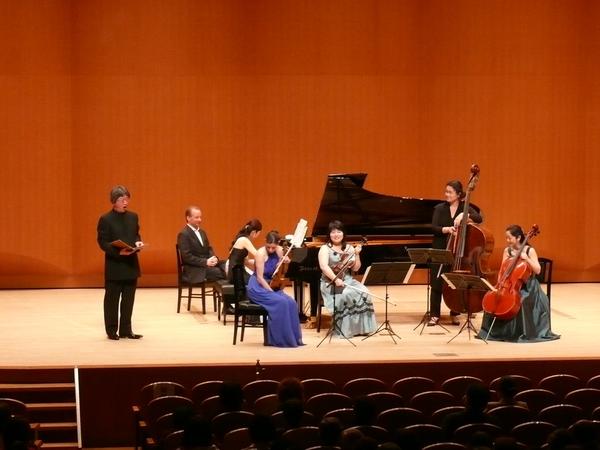 舞台で、ドレスとスーツを着た男女が、弦楽器とピアノを演奏し、その横で、男性が歌ってている写真