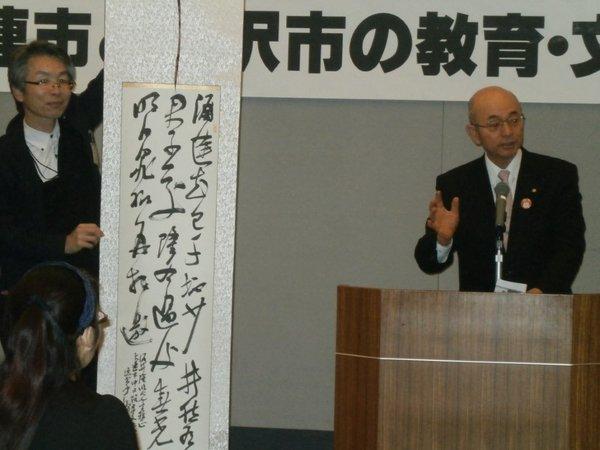 演台に立ち話をしている市長と隣で草書で書かれた書の掛軸を持っている男性の写真