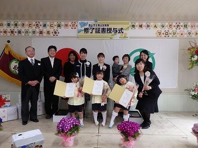 修了証書授与式の横断幕の下で卒園生3名と保護者及び関係者が一緒に写っている集合写真