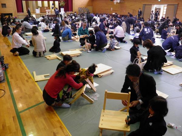 体育館一杯にシートを広げ、大人と子どもが一緒に木材を使って椅子を組み立てて作成しており、沢山の人が参加して作成している様子の写真