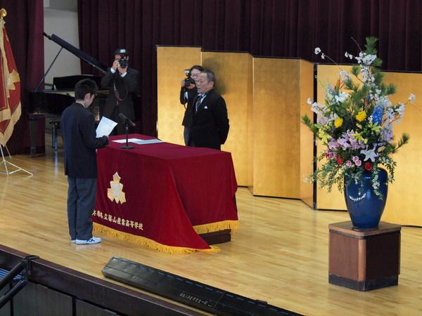 赤のテーブルクロスの演台の前に立っている礼服を着た男性を前に代表生徒が式辞を述べている写真