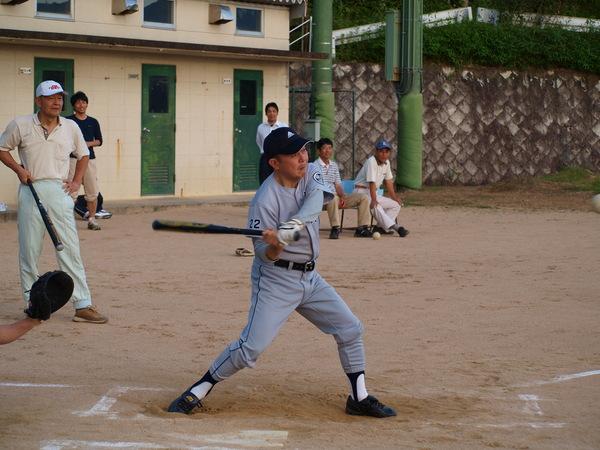 青い帽子をかぶり、グレーのユニフォームを着た足立議長がバッターボックスに立って、ボールを打とうと、バットを後ろに振り上げている様子の写真