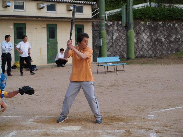 オレンジ色のポロシャツにグレーのジャージを着た小山部長がバッターボックスに立って、バットを握って構えている様子の写真