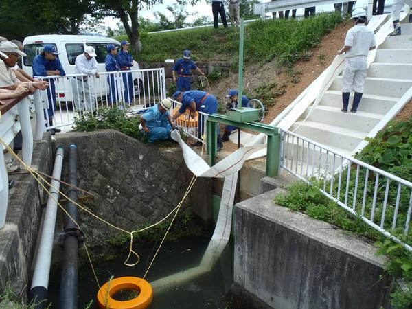 水門口の上で排水ポンプが設置され機械にホースの取り付けをしているのを関係者が集まって見ている様子の写真