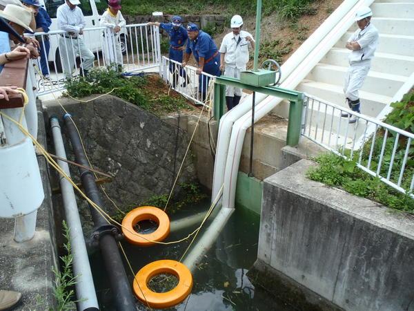 川の水門の入り口前に二つの黄色い浮き輪のようなものが落とされて、二つのホースから汲み上げられた水がポースを伝わって排出されており、消防関係者が見ている様子の写真