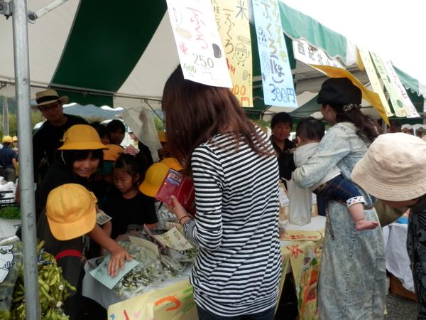 テントの中で子供達が販売していて赤い財布の女性が品物をみてその隣には赤ちゃんを抱えたお母さんが品物を手にもっている写真