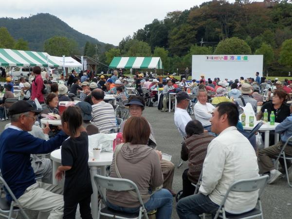 飲食コーナーで沢山の人が丸い白いテーブルの上にお茶や荷物をおきパイプ椅子に座り笑顔で談笑している写真