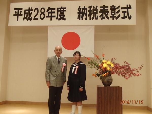平成28年度 納税表彰式の横断幕の前で市長と細見 佳世さんが綺麗な壇上花の横で二人で写っている写真