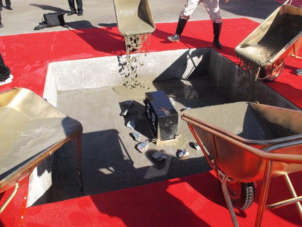 礎石と子ども達の夢が書かれてた石が置かれている四角に囲まれた枠の中に、4台の一輪車に入れられたセメントが流し込まれている様子の写真