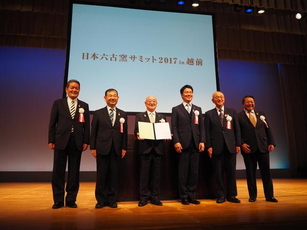 舞台上に市長とスーツを着た男性5人が並んでいる集合写真